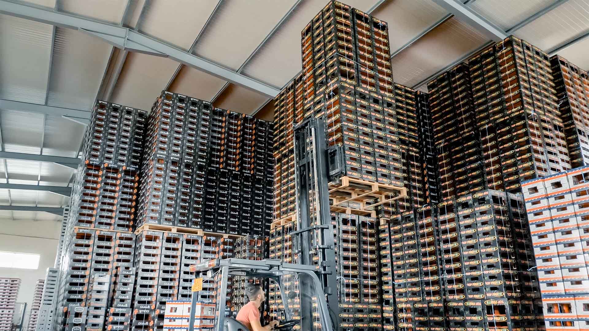 A forklift operator moving large pallets of packing materials stacking floor to ceiling in the corner of a warehouse