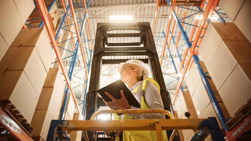 A warehouse worker on a forklift checking inventory with a note pad